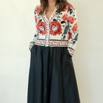 A woman modeling a long-sleeve button-up shirt with a vibrant red, black, and white floral pattern, paired with a black midi skirt and black strappy heels, against a plain off-white studio background.