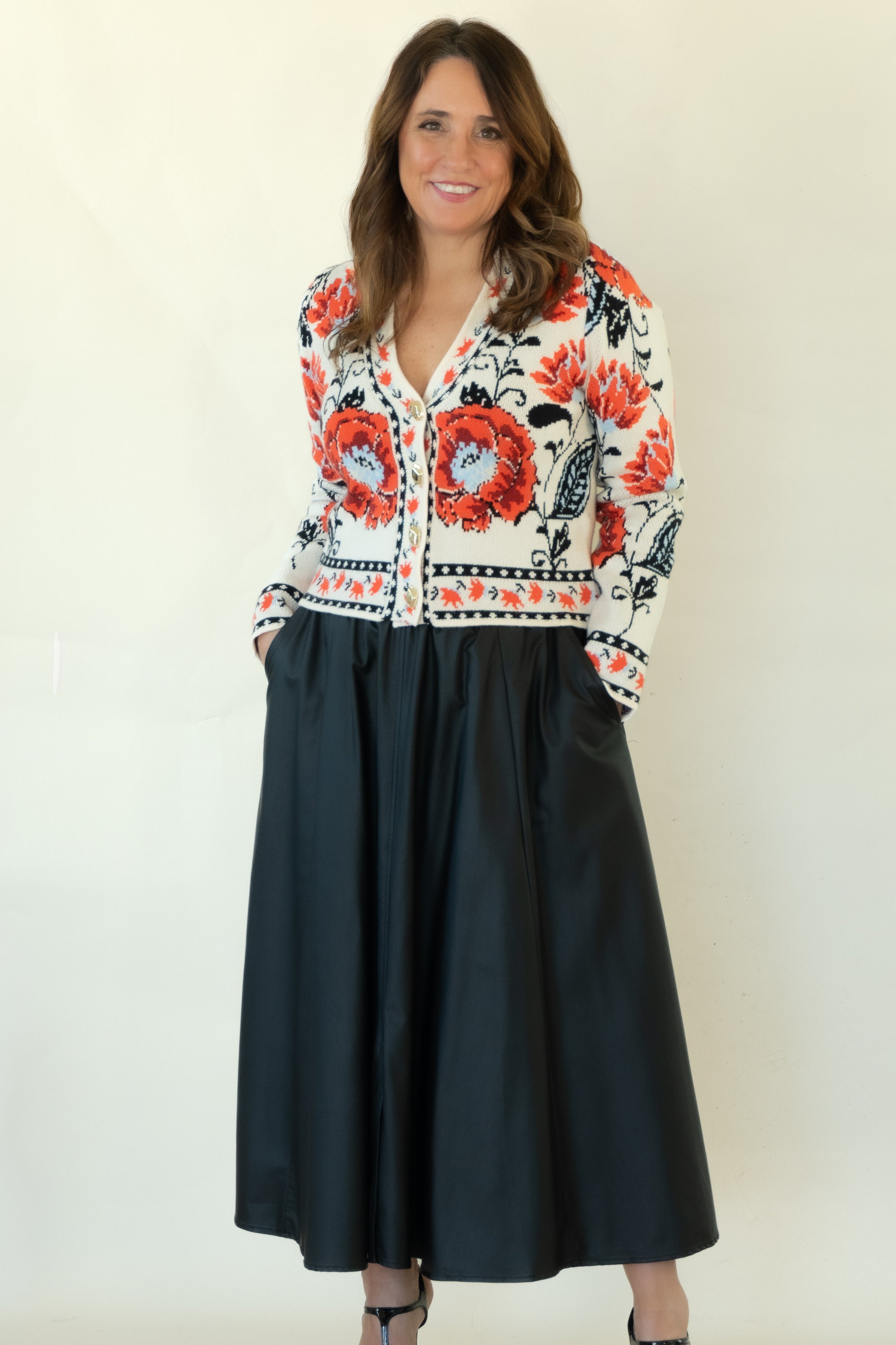 A woman modeling a long-sleeve button-up shirt with a vibrant red, black, and white floral pattern, paired with a black midi skirt and black strappy heels, against a plain off-white studio background.