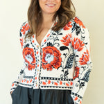 A woman modeling a long-sleeve button-up shirt with a white base and vibrant red, orange, and black floral patterns, paired with a dark charcoal gray midi skirt and black heels, against a plain cream background.