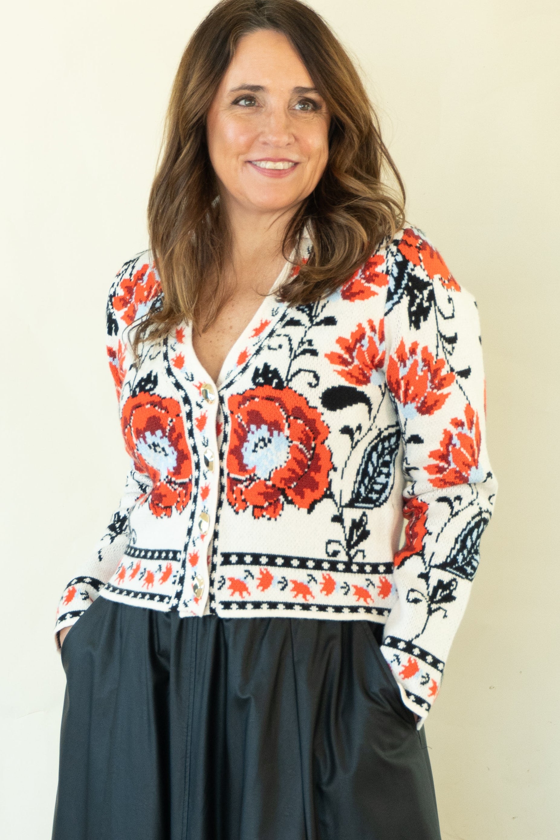 A woman modeling a long-sleeve button-up shirt with a white base and vibrant red, orange, and black floral patterns, paired with a dark charcoal gray midi skirt and black heels, against a plain cream background.