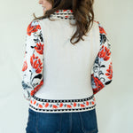 A woman shown from the back wearing a cream-colored knit cardigan with red, black, and white floral patterns on the sleeves and hem, paired with blue jeans against a plain off-white wall.