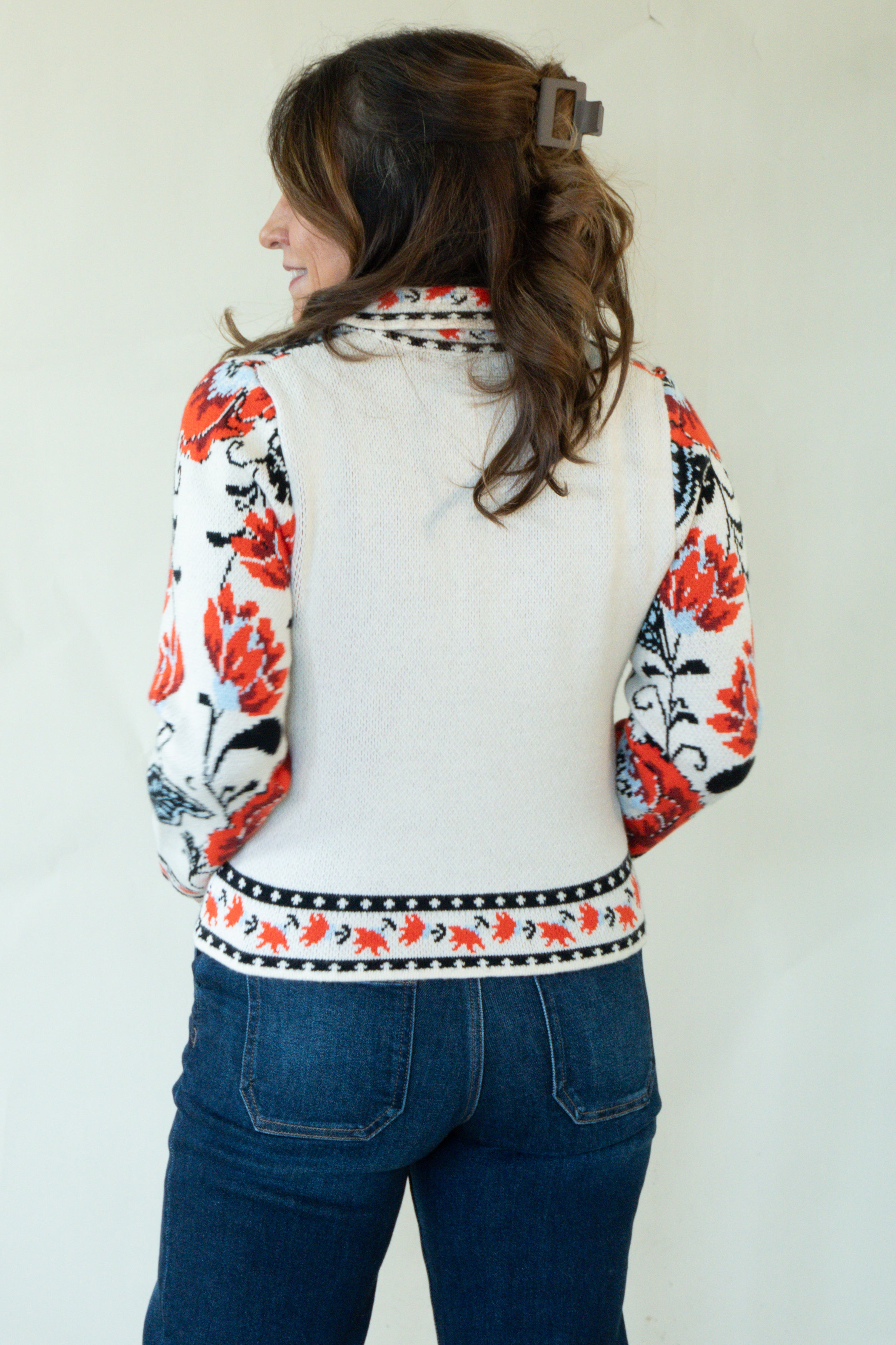 A woman shown from the back wearing a cream-colored knit cardigan with red, black, and white floral patterns on the sleeves and hem, paired with blue jeans against a plain off-white wall.