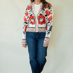 A woman modeling a white cardigan with a vibrant red and black floral pattern, paired with blue jeans and tan boots, standing against a plain off-white background.