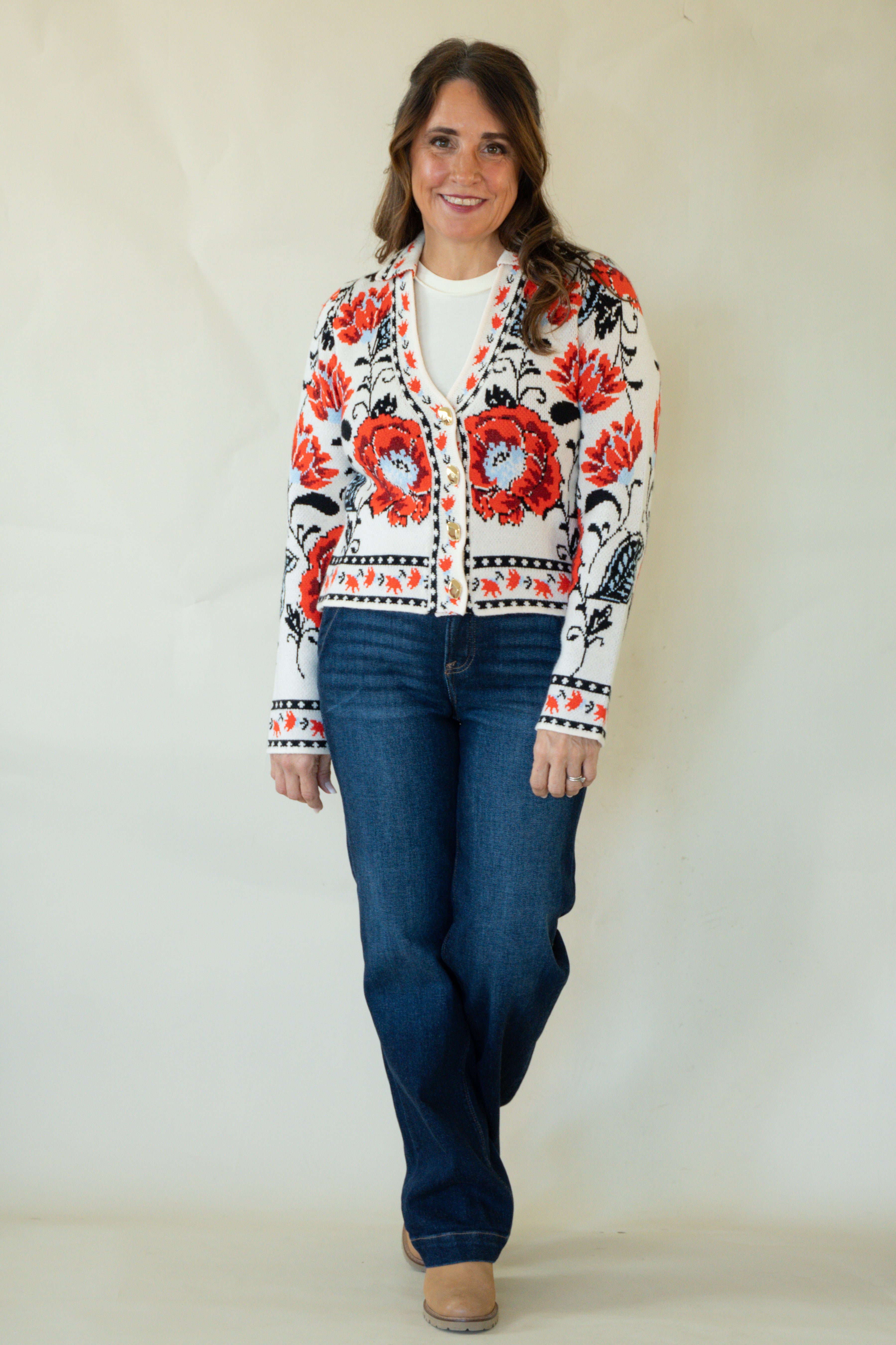 A woman modeling a white cardigan with a vibrant red and black floral pattern, paired with blue jeans and tan boots, standing against a plain off-white background.