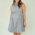 A woman modeling a sleeveless high-neck A-line dress with a blue, white, and beige gingham check pattern, standing against a plain cream-colored background.