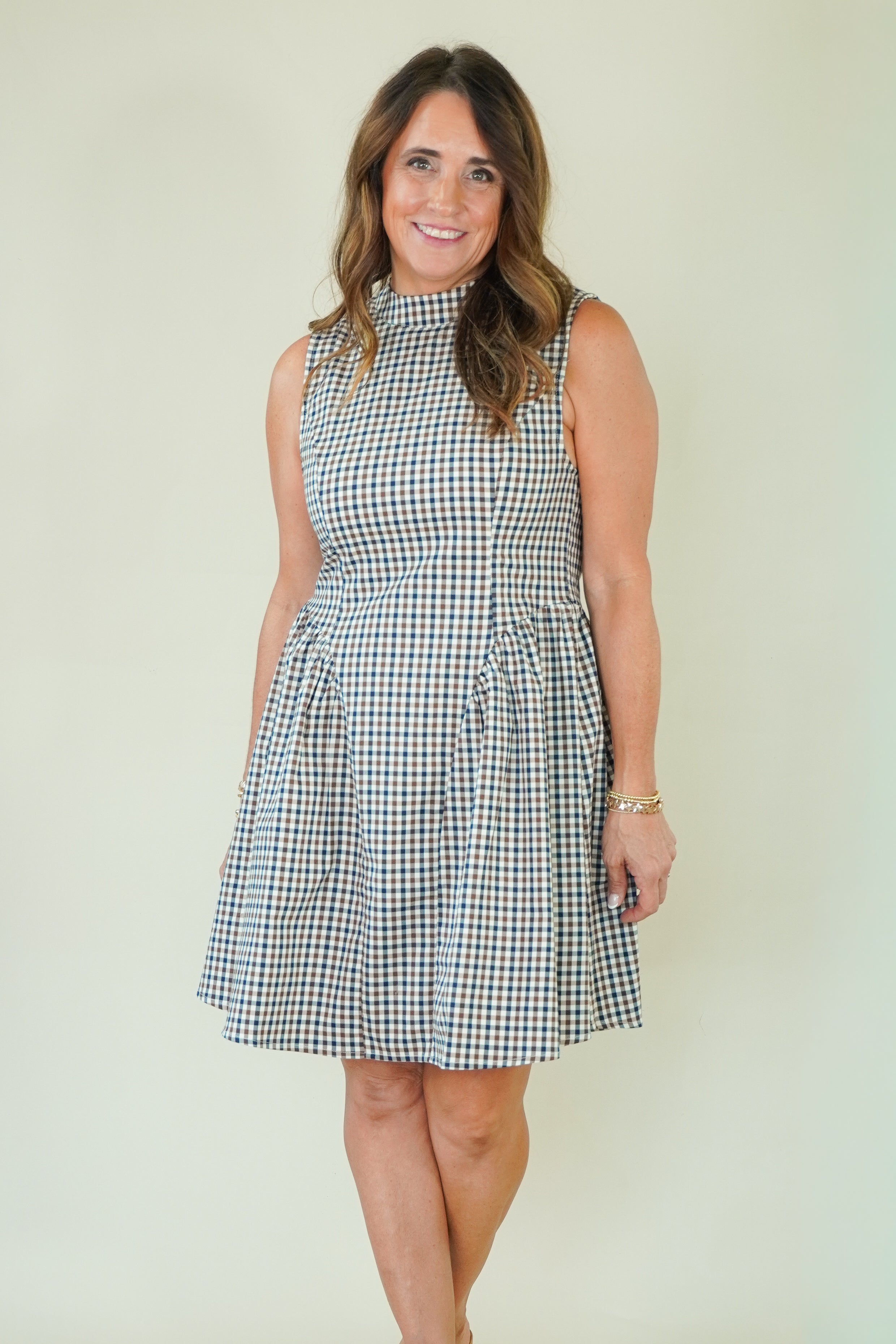 A woman modeling a sleeveless high-neck A-line dress with a blue, white, and beige gingham check pattern, standing against a plain cream-colored background.