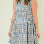 A woman modeling a sleeveless high-neck A-line dress with a blue, white, and beige gingham check pattern, standing against a plain cream-colored background.