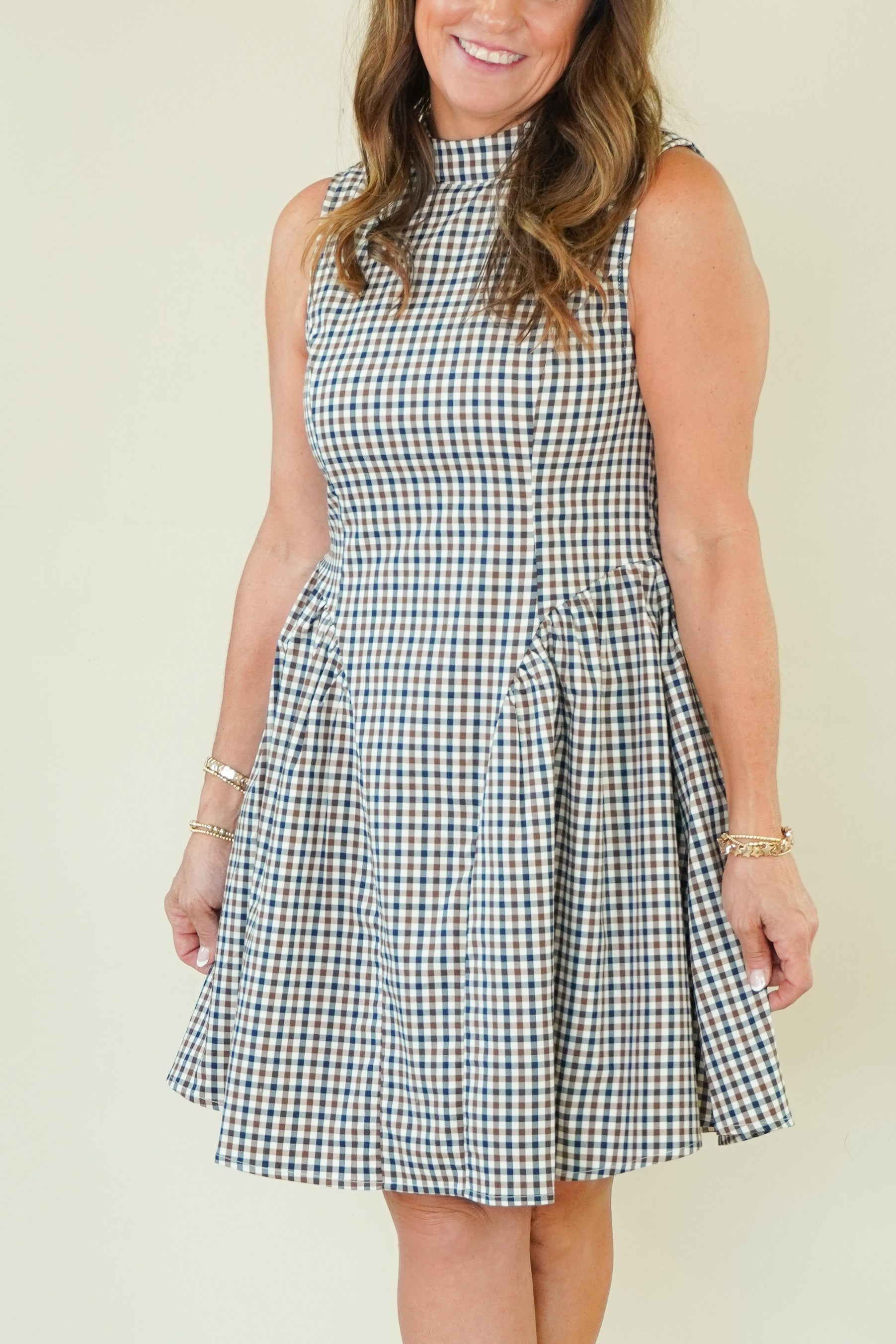 A woman modeling a sleeveless high-neck A-line dress with a blue, white, and beige gingham check pattern, standing against a plain cream-colored background.