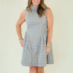 A woman modeling a sleeveless high-neck A-line dress with a blue, white, and beige gingham check pattern, standing against a plain cream-colored background.