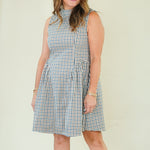 A woman modeling a sleeveless high-neck A-line dress with a blue, white, and beige gingham check pattern, standing against a plain cream-colored background.
