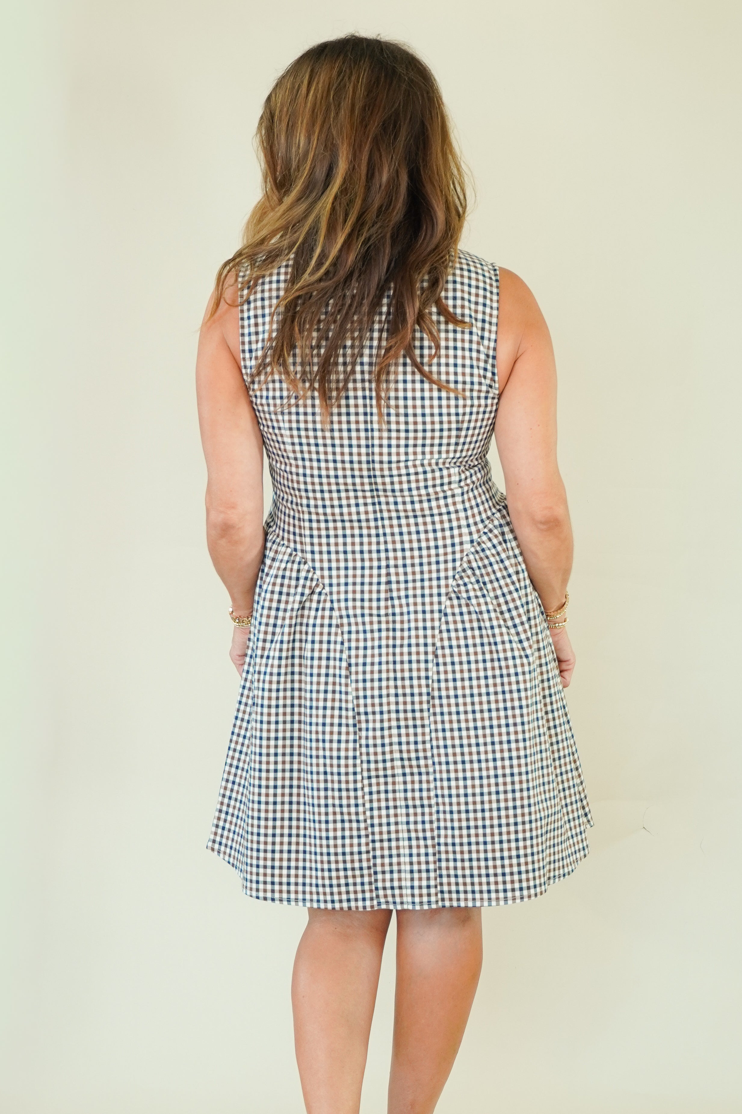 A woman modeling a sleeveless high-neck A-line dress with a blue, white, and beige gingham check pattern, standing against a plain cream-colored background.