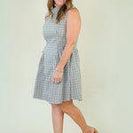 A woman modeling a sleeveless, knee-length gingham dress with a tie waist, paired with brown clogs, against a plain light beige studio background.