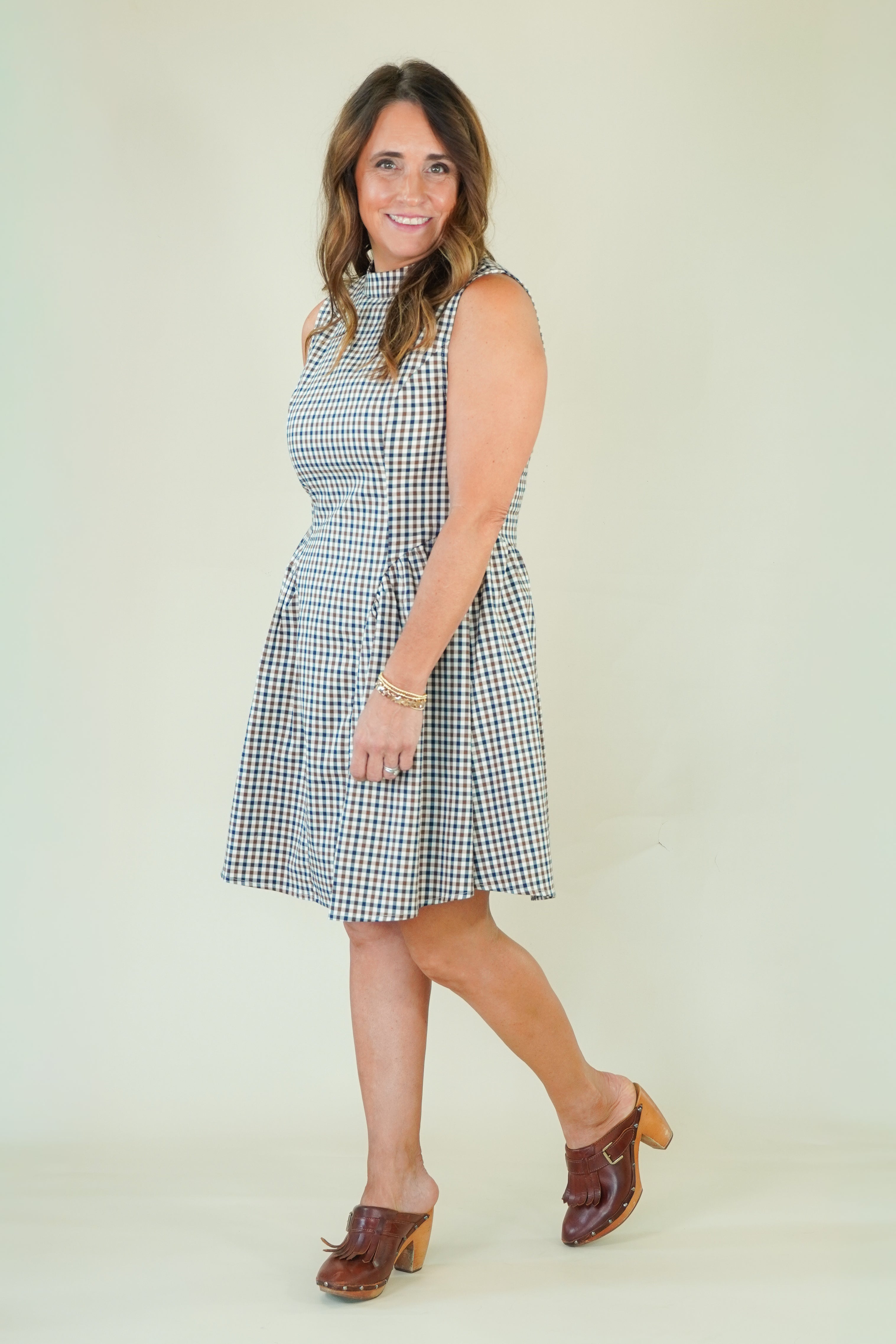 A woman modeling a sleeveless, knee-length gingham dress with a tie waist, paired with brown clogs, against a plain light beige studio background.