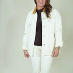 A woman modeling a white jacket and matching white pants over a dark brown turtleneck sweater, standing against a plain light beige studio backdrop.