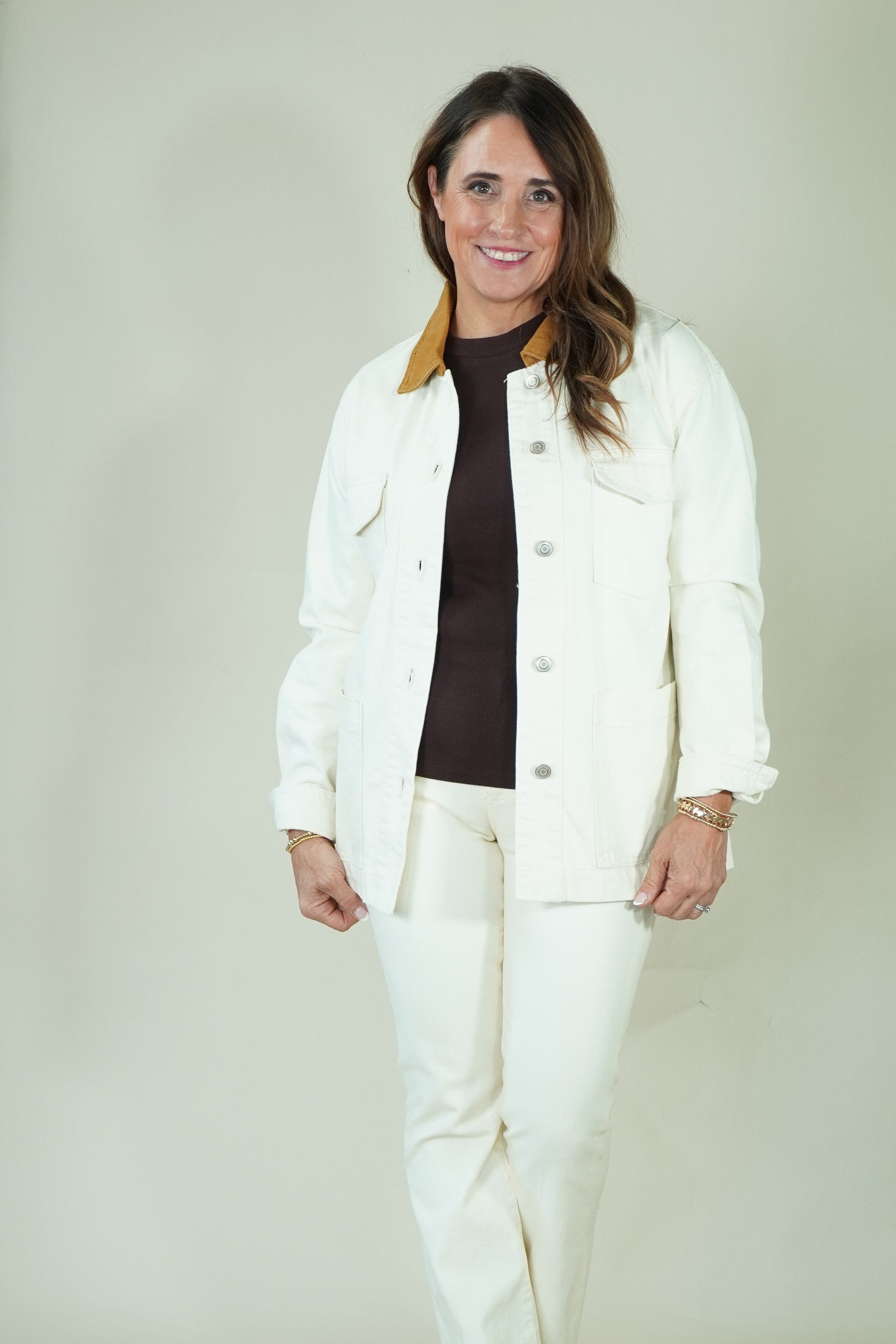 A woman modeling a white jacket and matching white pants over a dark brown turtleneck sweater, standing against a plain light beige studio backdrop.