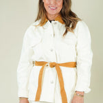 A woman modeling a white two-piece outfit consisting of a long-sleeve button-front jacket with a tan belt and matching white trousers, photographed against a plain light beige studio background.