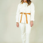 A woman modeling a white two-piece set consisting of a long-sleeve collared shirt with a tan belt and matching white pants, photographed against a plain light beige studio background.