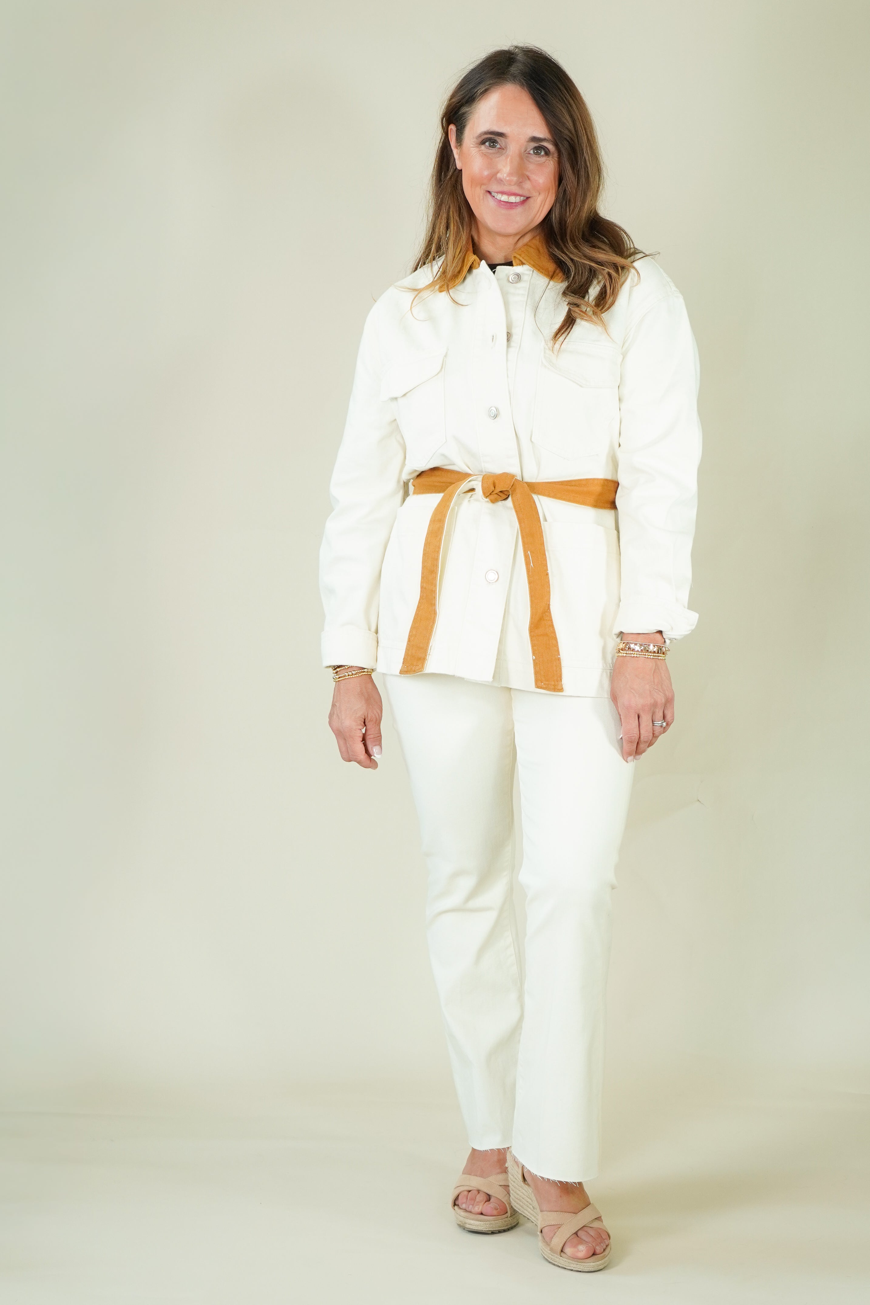 A woman modeling a white two-piece set consisting of a long-sleeve collared shirt with a tan belt and matching white pants, photographed against a plain light beige studio background.