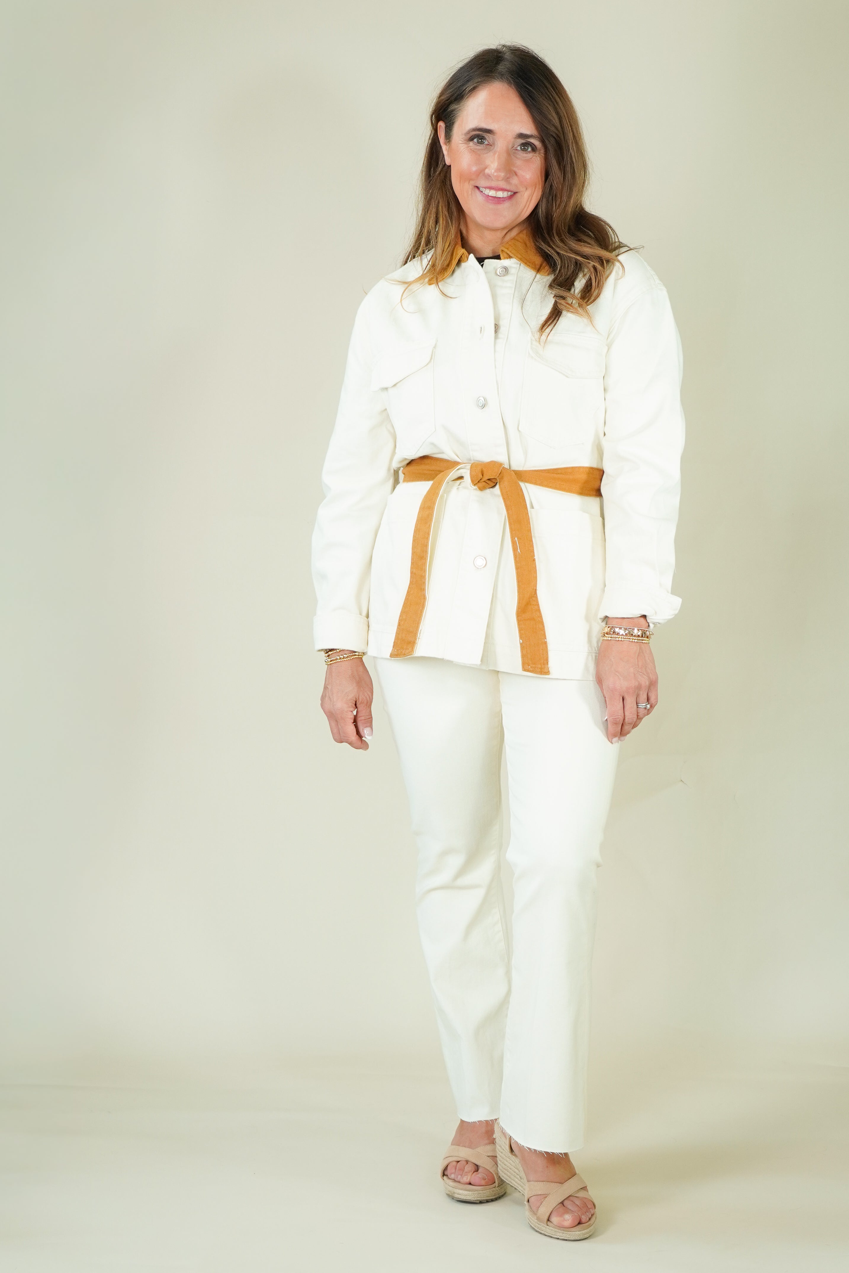 A woman modeling a white two-piece set consisting of a long-sleeve collared shirt with a tan belt and matching white pants, photographed against a plain light beige studio background.