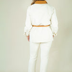 A woman viewed from behind wearing a white long-sleeve belted jacket with a tan collar and matching white pants, standing against a plain light beige studio background.