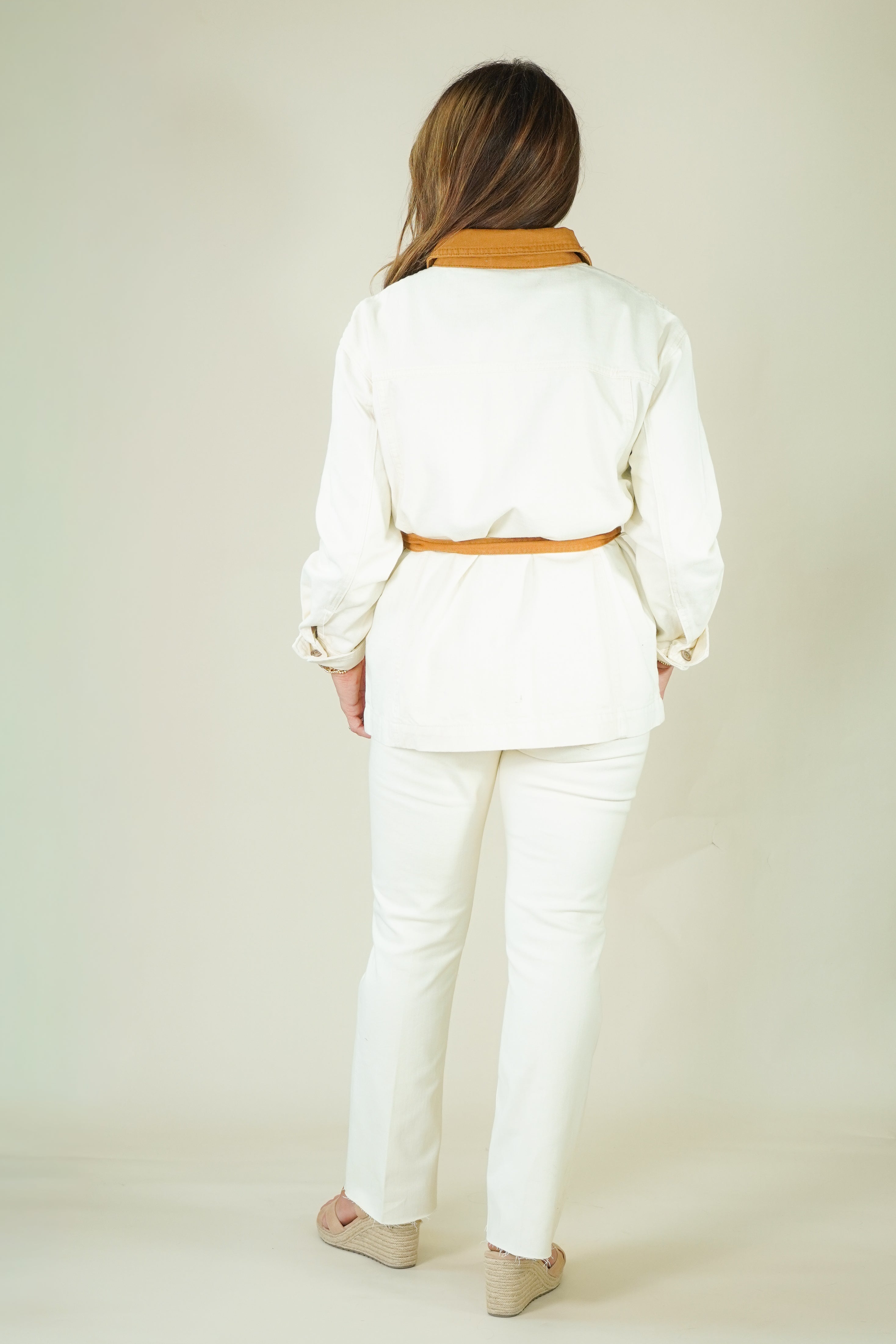 A woman viewed from behind wearing a white long-sleeve belted jacket with a tan collar and matching white pants, standing against a plain light beige studio background.