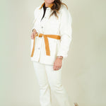 A woman modeling a white two-piece suit with a tan belt and matching tan collar detail, paired with beige wedge sandals, against a plain light beige studio background.