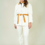 A woman modeling a white two-piece outfit consisting of a long-sleeve button-front jacket with a wide tan belt and matching white trousers, paired with tan heeled sandals against a plain cream studio background.