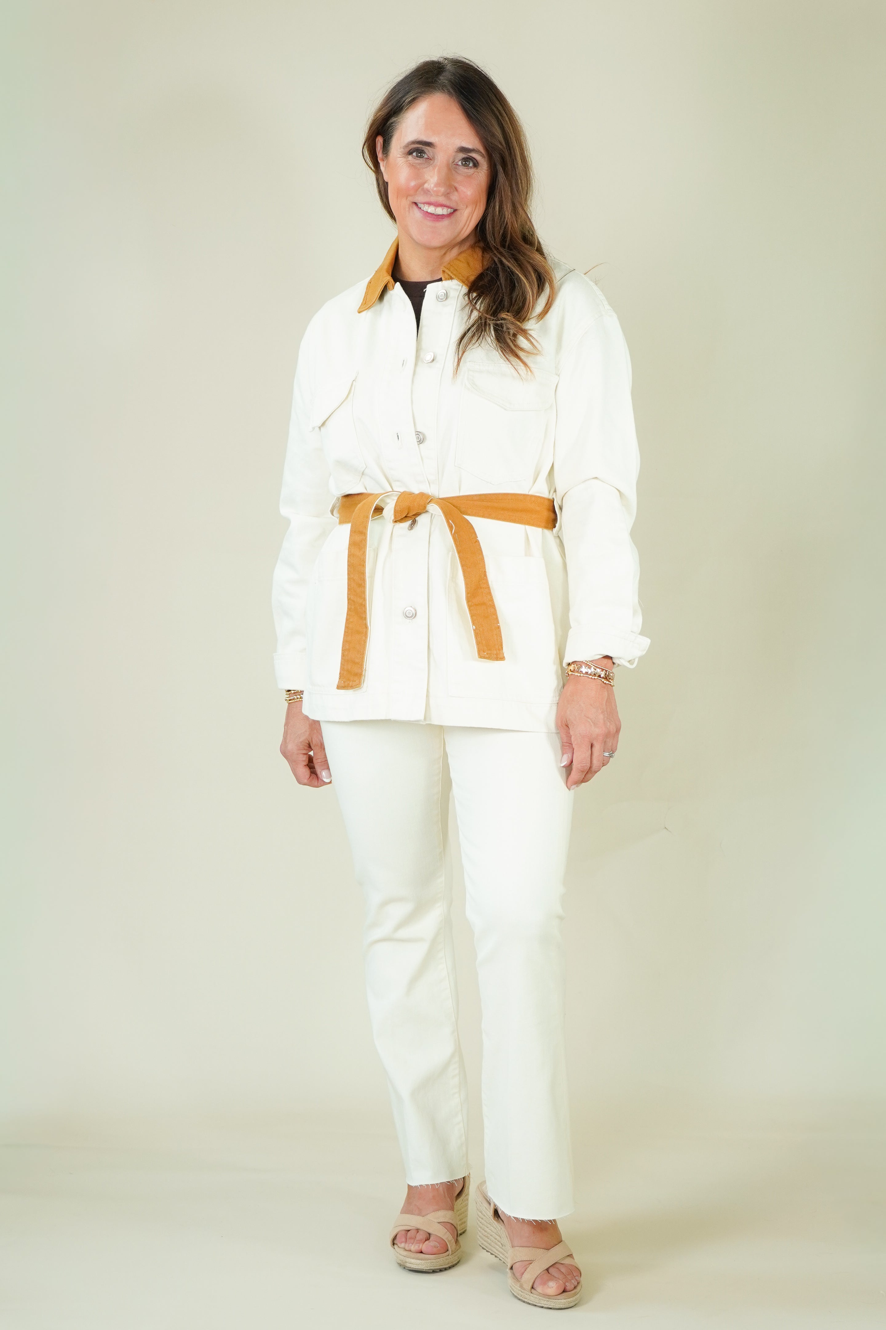 A woman modeling a white two-piece outfit consisting of a long-sleeve button-front jacket with a wide tan belt and matching white trousers, paired with tan heeled sandals against a plain cream studio background.