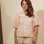 Woman standing in a room wearing a patterned shirt and shorts, with a mirror reflecting part of the room.