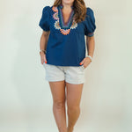 A woman modeling a navy blue short-sleeve blouse with colorful floral embroidery around the V-neck, paired with light beige shorts and brown sandals, standing against a plain off-white studio background.