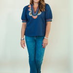 A woman modeling a navy blue short-sleeve blouse with colorful floral embroidery around the V-neck, paired with blue jeans and brown slide sandals, standing against a plain off-white studio background.
