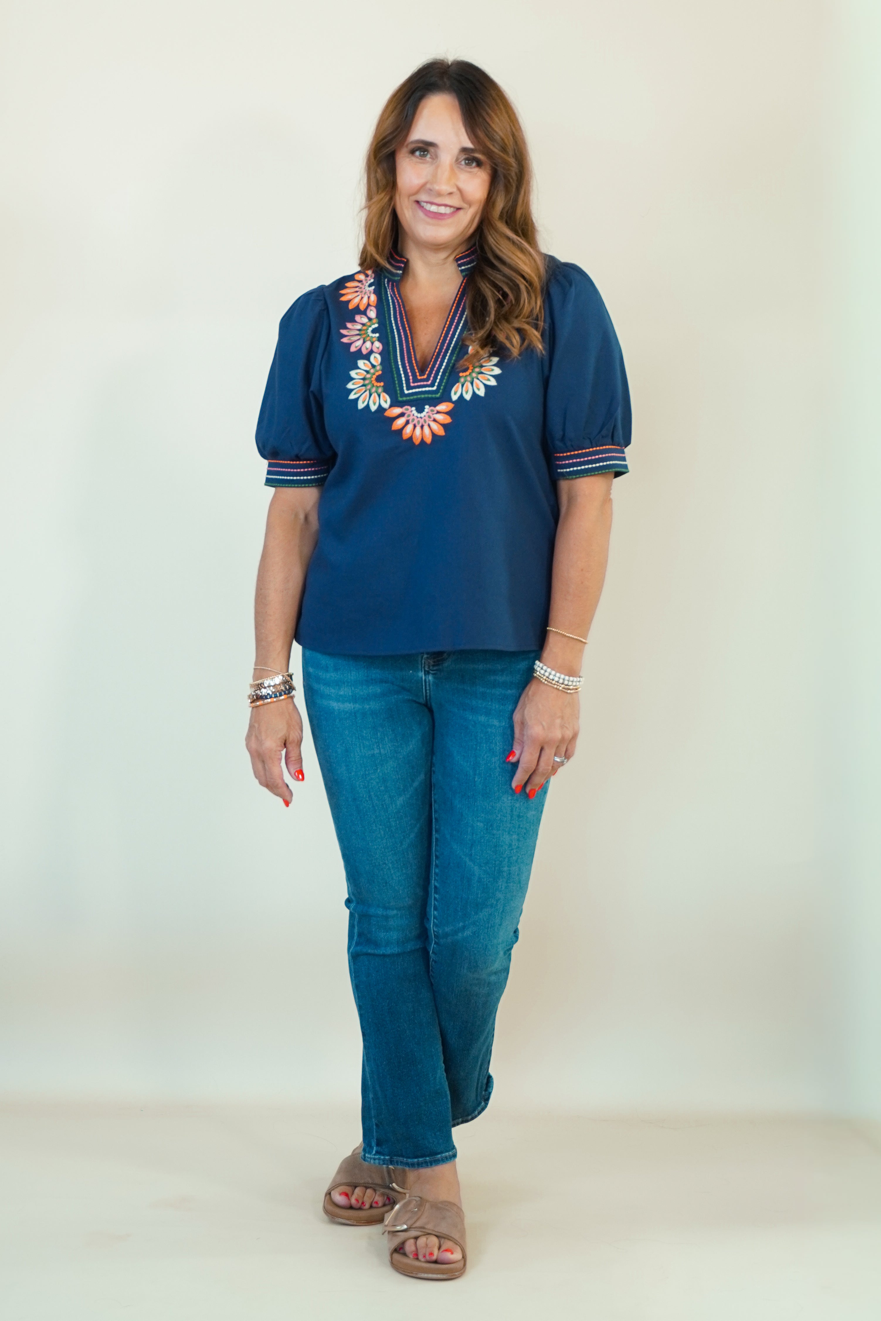 A woman modeling a navy blue short-sleeve blouse with colorful floral embroidery around the V-neck, paired with blue jeans and brown slide sandals, standing against a plain off-white studio background.