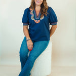 A woman wearing a navy blue short-sleeve blouse with colorful floral embroidery around the V-neck, paired with blue jeans and brown slide sandals, sitting on a white textured stool against a plain off-white studio background.