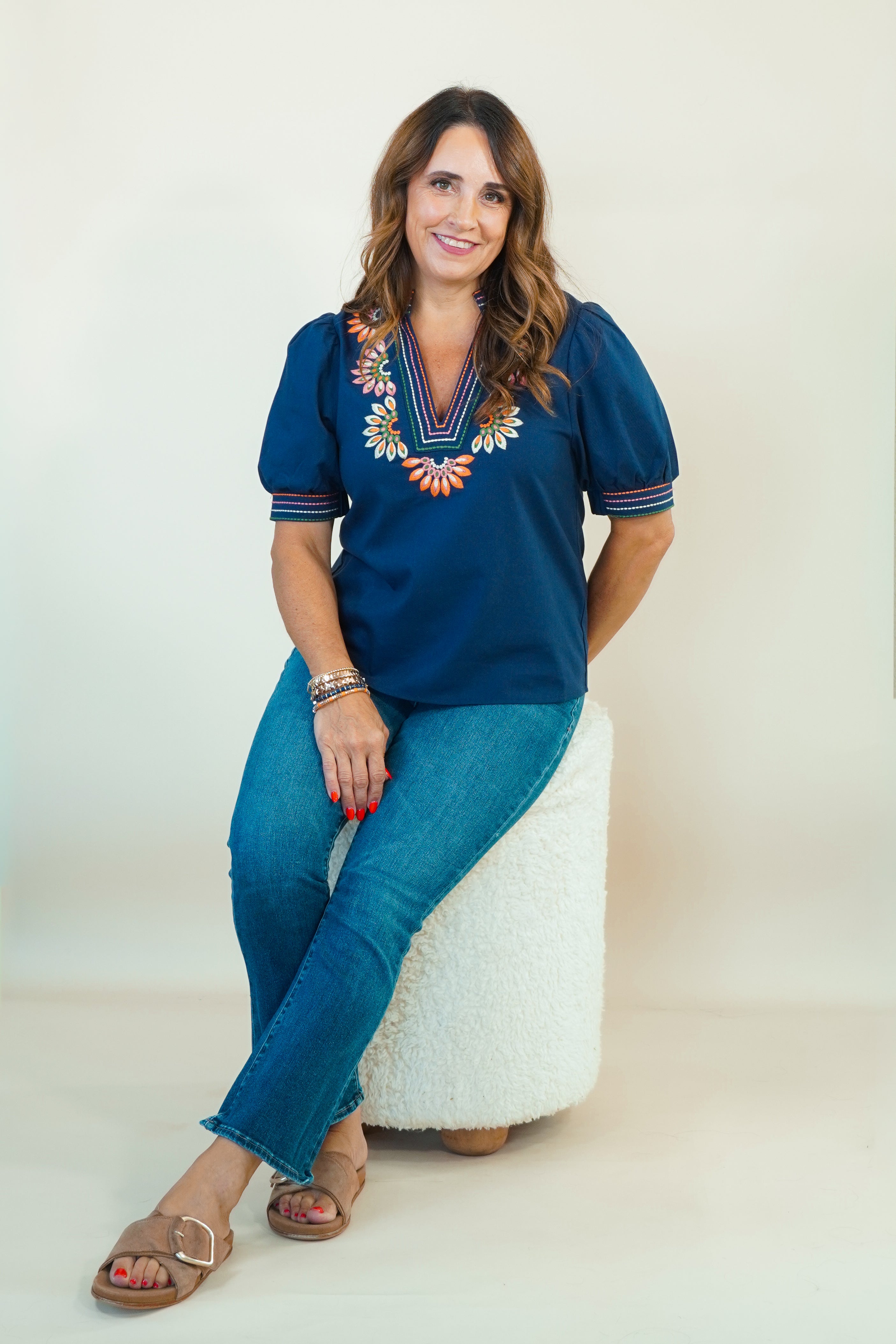 A woman wearing a navy blue short-sleeve blouse with colorful floral embroidery around the V-neck, paired with blue jeans and brown slide sandals, sitting on a white textured stool against a plain off-white studio background.