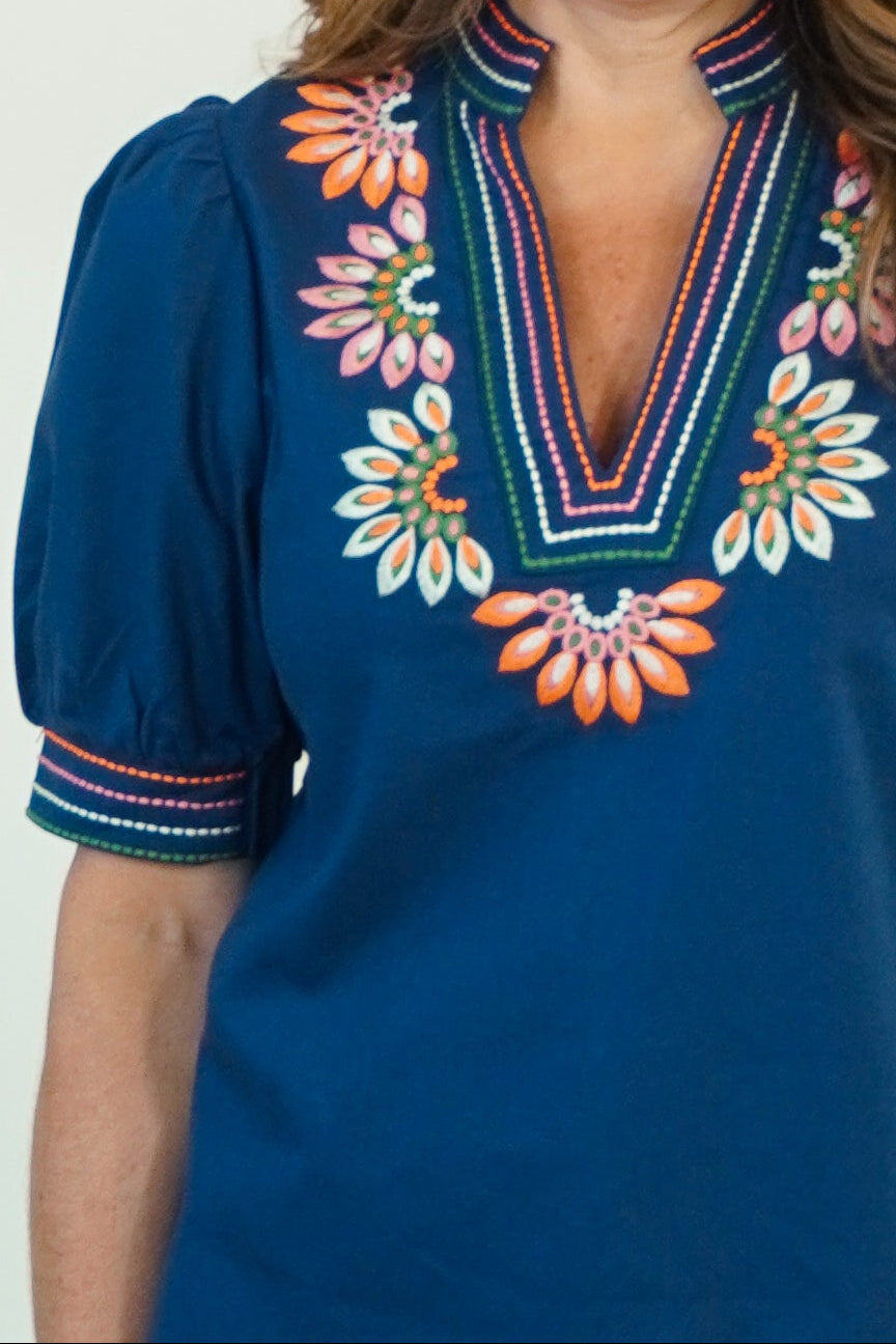 A close-up of a woman wearing a royal blue short-sleeve blouse with colorful embroidered floral details around the V-neck and sleeve cuffs.