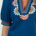 A close-up of a woman wearing a royal blue short-sleeve blouse with colorful embroidered floral details around the V-neck and sleeve cuffs.
