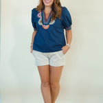A woman modeling a navy blue short-sleeve blouse with colorful floral embroidery around the V-neck, paired with white shorts and brown sandals, standing against a plain off-white studio background.