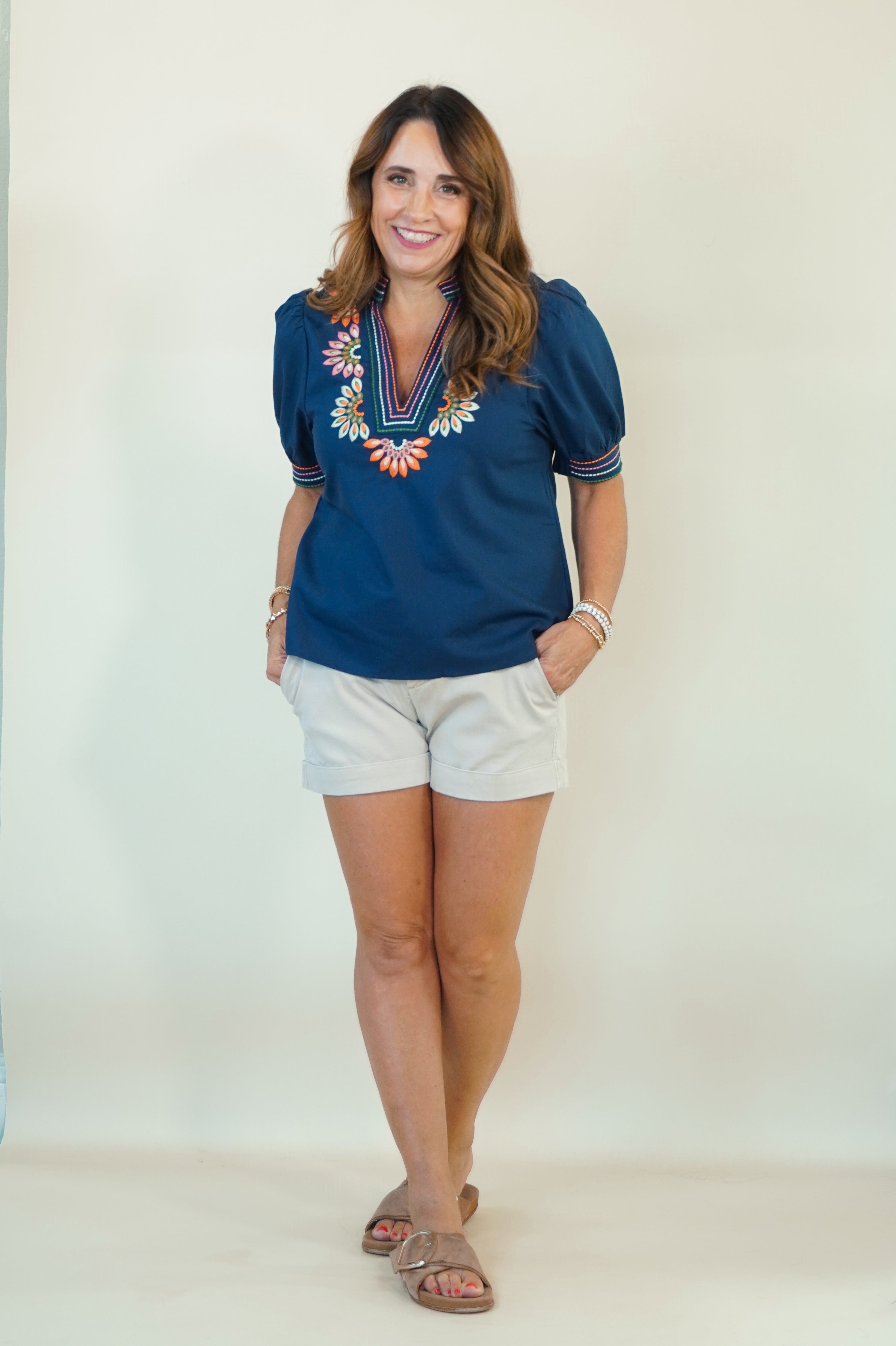 A woman modeling a navy blue short-sleeve blouse with colorful floral embroidery around the V-neck, paired with white shorts and brown sandals, standing against a plain off-white studio background.