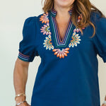 A woman modeling a navy blue short-sleeve blouse with colorful floral embroidery around the V-neck and sleeve cuffs, paired with white shorts and tan sandals, against a plain light beige studio background.