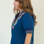 A woman wearing a navy blue short-sleeve blouse with colorful embroidered trim on the neckline and cuffs, paired with white shorts and brown slide sandals, shown in profile against a plain light background.