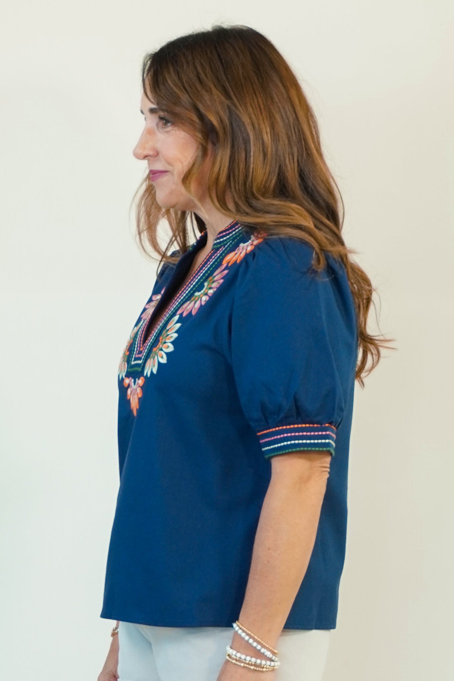 A woman wearing a navy blue short-sleeve blouse with colorful embroidered trim on the neckline and cuffs, paired with white shorts and brown slide sandals, shown in profile against a plain light background.