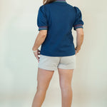 A woman shown from the back wearing a navy blue short-sleeve polo shirt with striped trim on the collar and cuffs, paired with light beige shorts and brown sandals, against a plain off-white background.