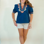 A woman modeling a navy blue short-sleeve top with colorful floral embroidery around the V-neck and sleeve cuffs, paired with white shorts, against a plain light background.