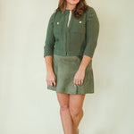 A woman modeling a sage green ribbed knit cardigan and matching corduroy mini skirt set against a plain cream background.