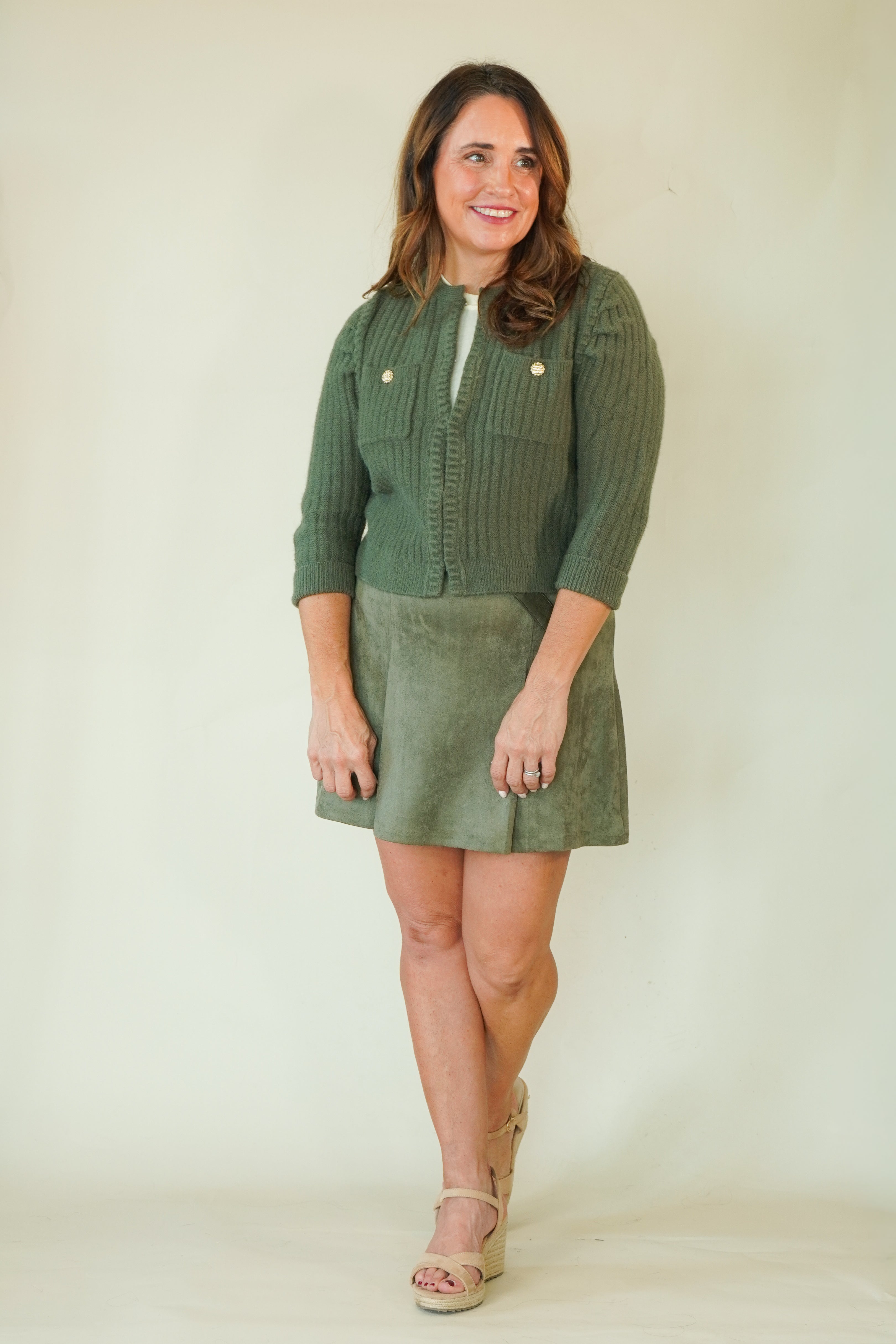A woman modeling a sage green ribbed knit cardigan and matching corduroy mini skirt set against a plain cream background.