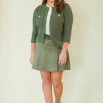 A woman modeling a matching olive green knit cardigan and skirt set with a white top and beige wedge sandals, standing against a plain cream background.