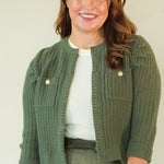 A woman modeling a coordinated outfit consisting of an olive green ribbed knit cardigan, a matching olive green mini skirt, a white top, and beige strappy sandals, set against a plain cream studio background.