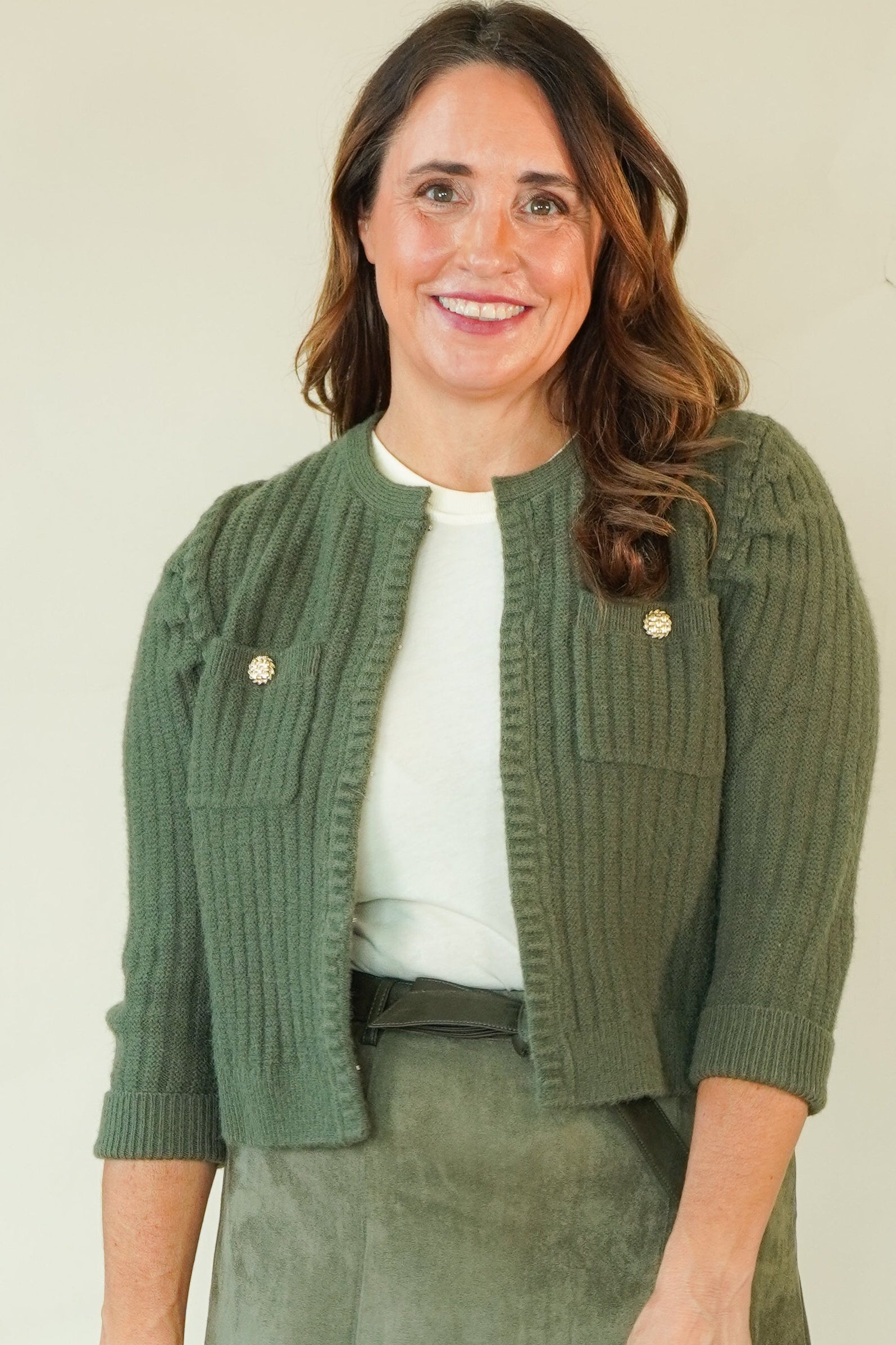 A woman modeling a coordinated outfit consisting of an olive green ribbed knit cardigan, a matching olive green mini skirt, a white top, and beige strappy sandals, set against a plain cream studio background.