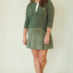 A woman modeling a two-piece olive green outfit consisting of a ribbed knit cardigan with gold buttons and a matching corduroy skirt, paired with beige platform sandals, against a plain cream background.