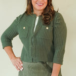 A woman modeling a matching olive green two-piece set consisting of a textured button-front cardigan and a corduroy mini skirt, paired with beige wedge sandals, against a plain cream background.